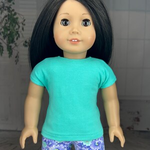 May include: A doll wearing a turquoise short-sleeved shirt and purple floral print shorts.