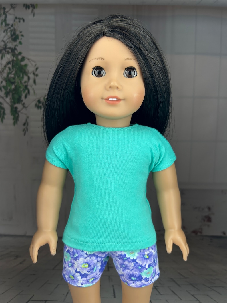 May include: A doll wearing a turquoise short-sleeved shirt and purple floral print shorts.