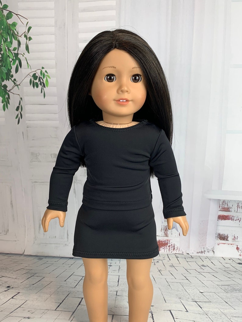 May include: A black long-sleeved dress with a round neckline and a skirt that comes just above the knee. The dress is being worn by a doll with long black hair and brown eyes.