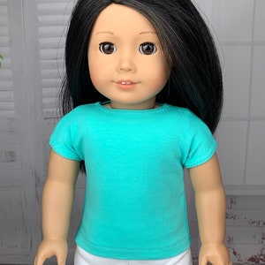 May include: A doll wearing a turquoise short-sleeved t-shirt and white pants.