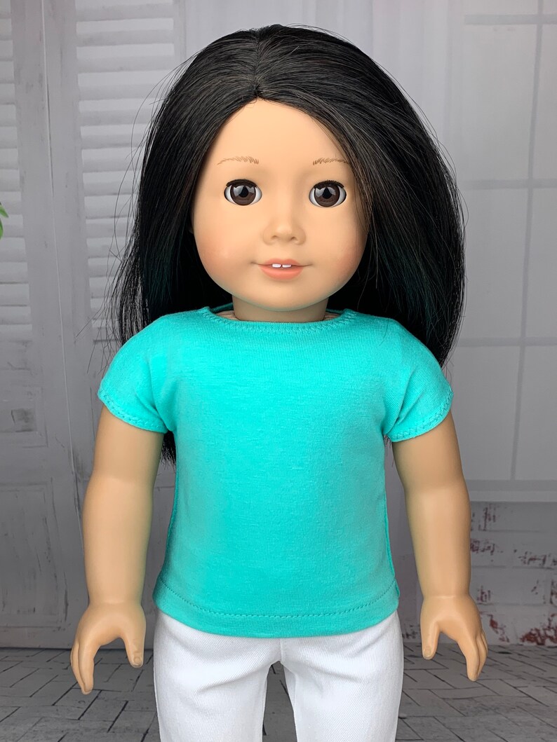 May include: A doll wearing a turquoise short-sleeved shirt and white pants.