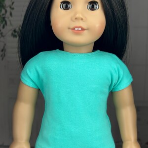 May include: A turquoise short-sleeved t-shirt for a doll. The doll is wearing a floral print pair of pants.