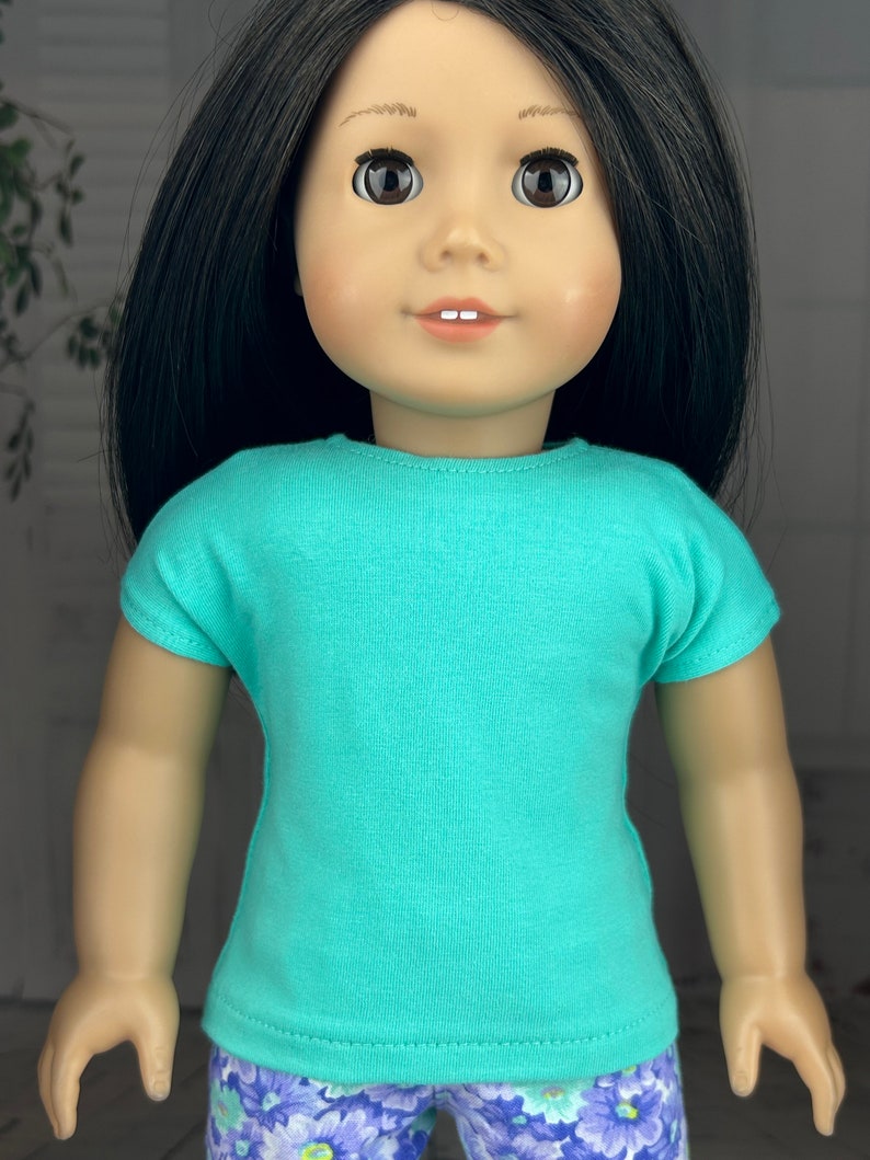 May include: A turquoise short-sleeved t-shirt for a doll. The doll is wearing a floral print pair of pants.