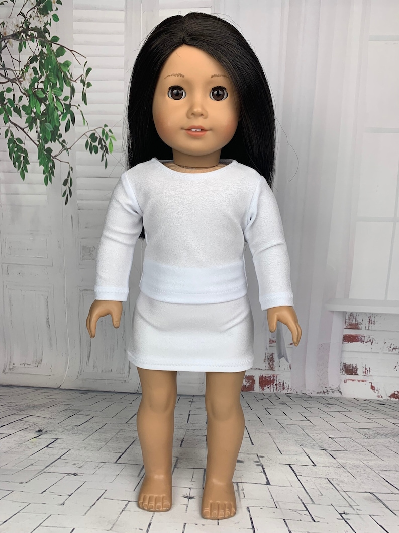 May include: A white long-sleeved shirt and a white skirt outfit for a doll. The doll has long black hair and brown eyes.