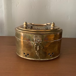 Könnte beinhalten: Eine runde Messingdose mit einem Scharnierdeckel und einem dekorativen Verschluss. Die Dose hat kleine Löcher an den Seiten und einen Griff oben.