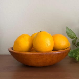 May include: A round, medium-brown wooden bowl filled with several bright yellow lemons. The bowl has a smooth, polished finish and a curved shape, holding the citrus fruit in a cluster. The lemons have a textured, vibrant yellow peel.