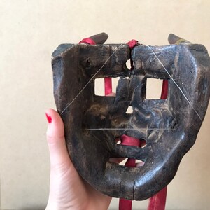 May include: A wooden mask with a carved face and holes for eyes and mouth. The mask is tied with red string and has a dark brown finish.