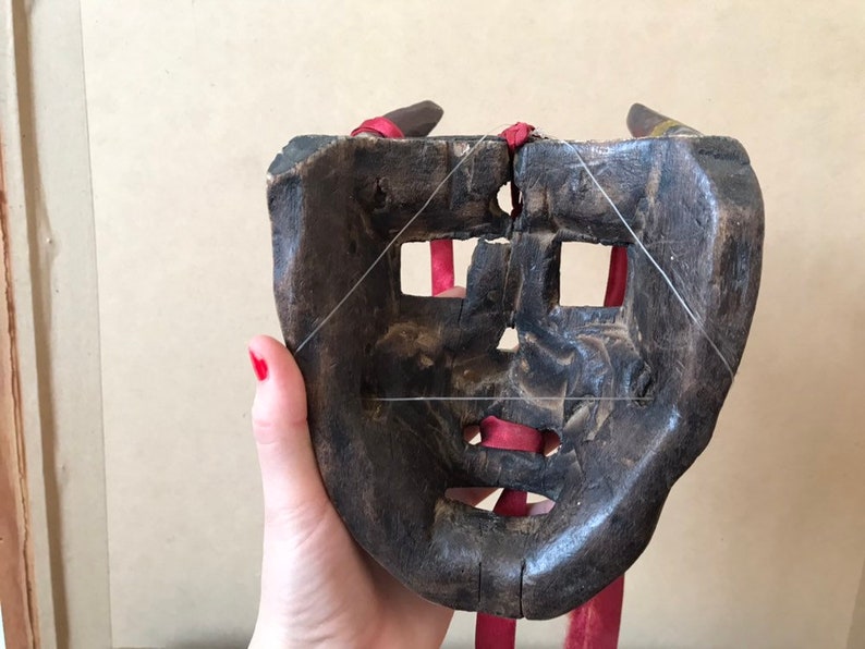 May include: A wooden mask with a carved face and holes for eyes and mouth. The mask is tied with red string and has a dark brown finish.