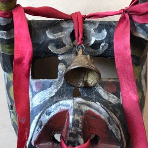 May include: A black wooden mask with red, white, and green paint. The mask has a bell attached to the top and is tied with a red ribbon.