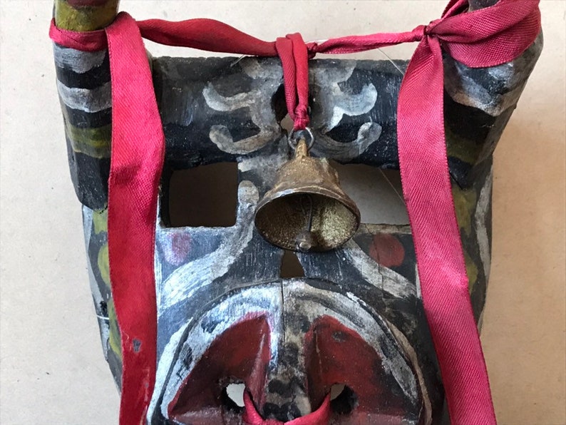 May include: A black wooden mask with red, white, and green paint. The mask has a bell attached to the top and is tied with a red ribbon.