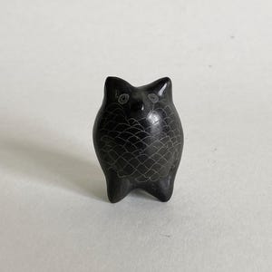 Puede incluir: Pequeña figura de búho negro con detalles grabados. El búho tiene un cuerpo redondeado, pies pequeños y ojos grandes. La superficie está texturizada con un patrón de escamas. Un objeto decorativo.