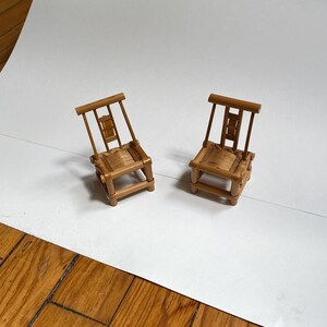 Puede incluir: Dos sillas de madera en miniatura con acabado marrón claro. Cada silla tiene un asiento rectangular, cuatro patas y un respaldo con un diseño decorativo. Las sillas están sobre una superficie blanca.