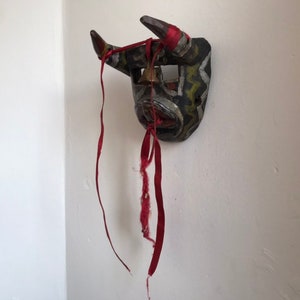 May include: A wooden bull mask with black, white, and green paint, and red ribbons attached. The mask has a bell hanging from the top.
