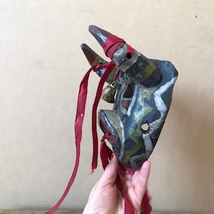 May include: A hand holds a wooden bull mask with red, black, white, and green paint. The mask has two horns and a bell hanging from the bottom. The mask is decorated with a red ribbon.