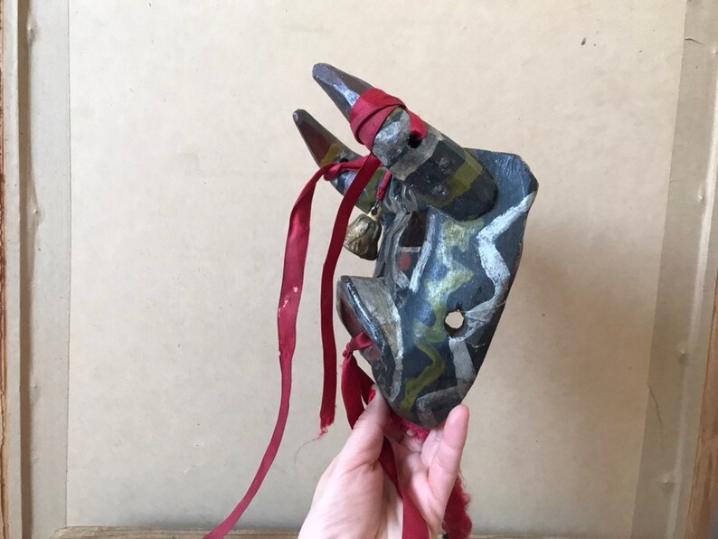 May include: A hand holds a wooden bull mask with red, black, white, and green paint. The mask has two horns and a bell hanging from the bottom. The mask is decorated with a red ribbon.