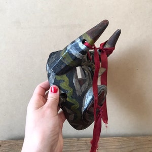 May include: A hand-carved wooden mask with horns, painted in black, green, white, and yellow. The mask has a red ribbon tied around the horns.