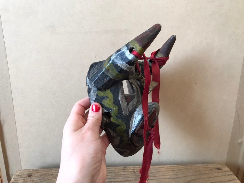 May include: A hand-carved wooden mask with horns, painted in black, green, white, and yellow. The mask has a red ribbon tied around the horns.