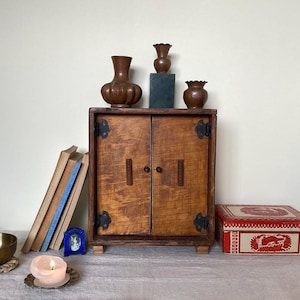 Puede incluir: Un armario de madera con dos puertas, una pila de libros y tres jarrones de cobre en la parte superior. El armario es marrón con un patrón de veta de madera. Los jarrones son de diferentes formas y tamaños. Los libros están apilados en el lado izquierdo del armario. El armario está sentado sobre una superficie marrón claro.