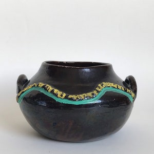 Puede incluir: Una olla de cerámica marrón oscuro con dos asas. La olla tiene un diseño pintado de verde y amarillo alrededor del borde.