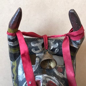 May include: A wooden mask with horns painted in black, white, green, and red. The horns are tied together with a red ribbon and a small bell hangs from the ribbon.