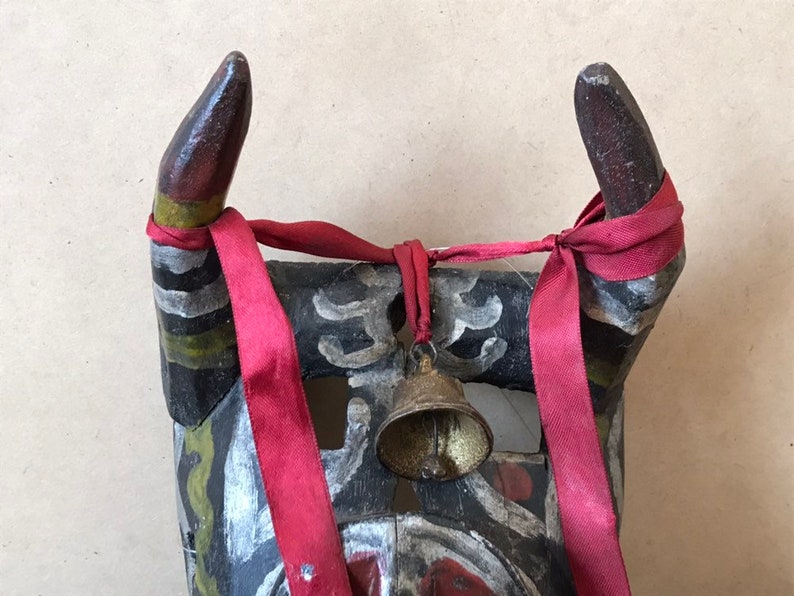 May include: A wooden mask with horns painted in black, white, green, and red. The horns are tied together with a red ribbon and a small bell hangs from the ribbon.