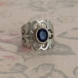 Puede incluir: Un anillo de plata con un diseño intrincado que presenta una piedra preciosa azul oscuro engastada en un bisel dorado.