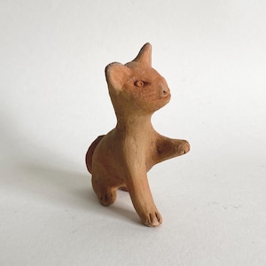 Puede incluir: Pequeña figura de gato de terracota hecha a mano. El gato está representado en una pose de caminar con una pata levantada. La escultura tiene un diseño simple y estilizado y un color marrón claro.