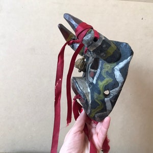 May include: A wooden bull mask with black, white, green, and red paint. The mask has two horns and a red ribbon attached to it.