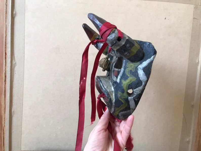 May include: A wooden bull mask with black, white, green, and red paint. The mask has two horns and a red ribbon attached to it.