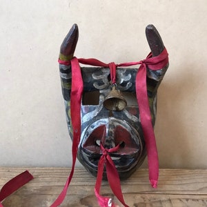 May include: A wooden mask with horns, painted in black, red, white, and green. The mask has a bell attached to the forehead and is tied with red ribbons.