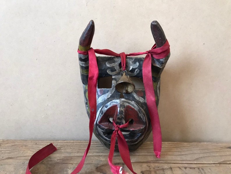 May include: A wooden mask with horns, painted in black, red, white, and green. The mask has a bell attached to the forehead and is tied with red ribbons.