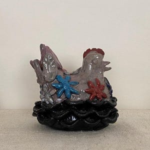 Puede incluir: Una figura de gallina de cerámica con cuerpo de color violeta claro brillante, con decoraciones en relieve en forma de flores azules y rojas. La gallina se asienta sobre una base negra ondulada y esmaltada que recuerda a un nido.