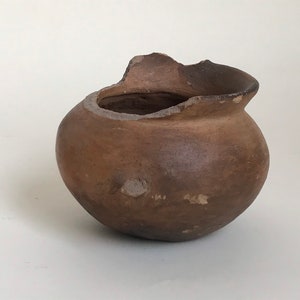 Puede incluir: Una pequeña olla de cerámica marrón con un borde astillado. La olla es redonda y tiene una superficie ligeramente irregular.
