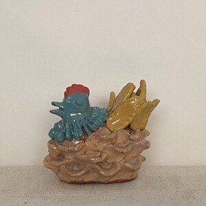 Puede incluir: Una figura de cerámica de un pollo azul con cresta roja sentado en un nido texturizado de color marrón claro. El pollo tiene una cola amarilla y un acabado brillante. La base del nido está esculpida con formas redondeadas superpuestas.