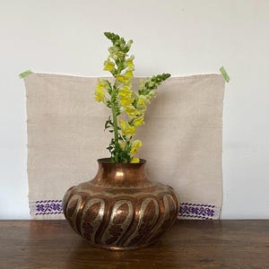 Könnte beinhalten: Eine Kupfervase mit einem verzierten Design enthält gelbe und grüne Blumen. Die Vase steht auf einer Holzoberfläche, mit einem beigen Tuch und lila gestickten Bordüren im Hintergrund. Das Tuch ist an der Wand befestigt.