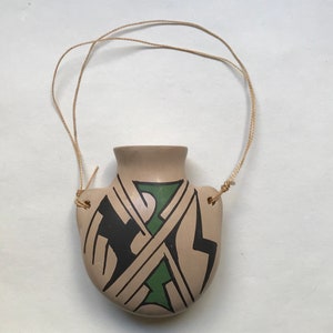 Puede incluir: Una pequeña maceta de cerámica de color beige con un diseño geométrico verde, negro y blanco. La maceta tiene una cuerda atada a ella, lo que permite colgarla.