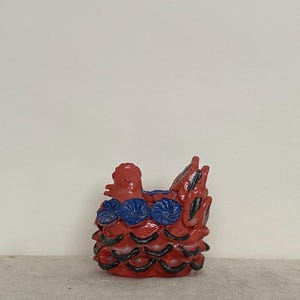 May include: A ceramic sculpture of a chicken with a glossy red finish, featuring three blue flower-like accents along the side and black painted details on the lower feathers. The figure has a raised head and a textured, layered feather design.