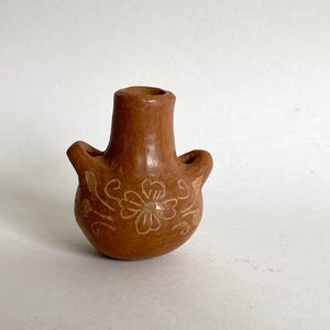 Puede incluir: Un pequeño jarrón de cerámica marrón con dos asas y un cuello estrecho. El jarrón presenta un diseño floral grabado en la superficie. El jarrón mide aproximadamente 10 cm de altura.