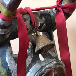 May include: A wooden mask with black, white, and green paint, decorated with red ribbons and a small brass bell.