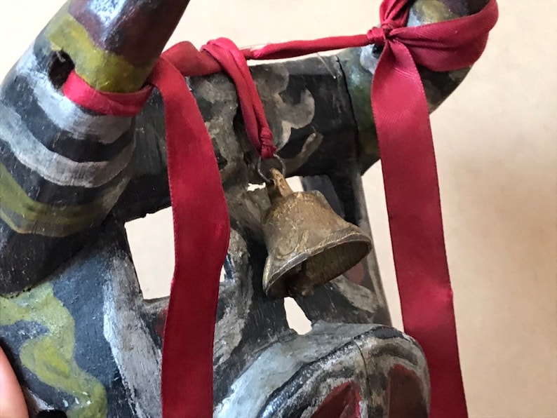 May include: A wooden mask with black, white, and green paint, decorated with red ribbons and a small brass bell.