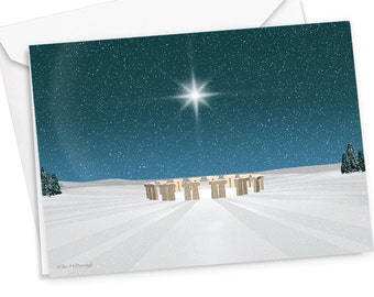 May include: A greeting card featuring an illustration of Stonehenge standing in a snow-covered landscape under a dark teal night sky. A bright, glowing star shines above the stone circle, and light snow falls across the scene. The artist name Ellen McDevitt is printed in the bottom left corner.