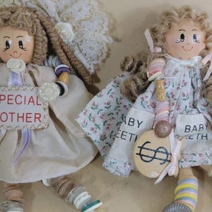 May include: Two fabric dolls with yarn hair and painted faces. The left doll wears a cream dress and holds a sign reading SPECIAL MOTHER. The right doll wears a floral dress with text reading BABY TEETH and holds a round wooden sign with a dollar sign.