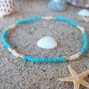May include: A turquoise and white beaded bracelet with gold accents. The bracelet is lying on a sandy surface with a seashell and a starfish.