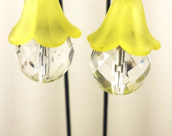 May include: A pair of earrings featuring a yellow flower-shaped top and a clear, faceted crystal orb. The earrings hang from a black cord and are suspended against a white background. The earrings are a unique accessory.