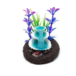 May include: A miniature blue fountain with three tiers, surrounded by green and purple plastic plants, on a brown base with small decorative shells.