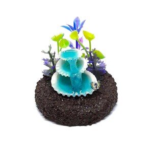 May include: A decorative miniature fountain with a cascading turquoise water effect. The fountain is set on a dark brown, textured base and adorned with colorful artificial flowers. A small butterfly embellishes the fountain.