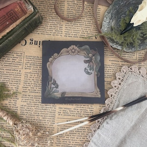 May include: A black and white decorative frame with a floral design. The frame has a blank space in the center for writing or drawing. The frame is surrounded by a dark background.