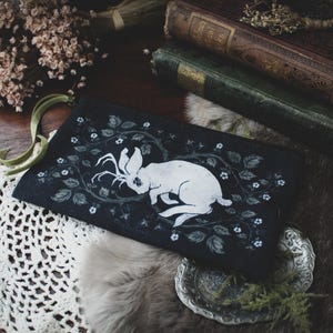 Puede incluir: Una bolsa de tela negra con una ilustración de un conejo blanco. El conejo tiene cuernos y está rodeado de diseños florales estilizados.