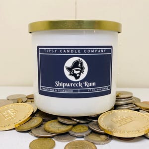 May include: A white candle with a gold lid, labeled "Shipwreck Rum" with a pirate logo. The candle is 17 oz and sits among a pile of coins, some gold and some silver.