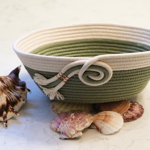 Puede incluir: Una cesta redonda de cuerda de dos tonos, verde y crema. La cesta tiene un asa de cuerda y borlas. Varias conchas marinas están dispuestas alrededor de la base de la cesta.
